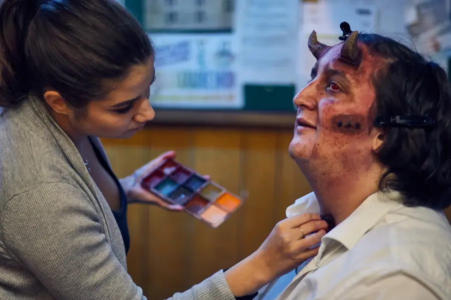 Szandra applying special-effects makeup to the neck of a model wearing devil horns, holding a makeup palette in her other hand